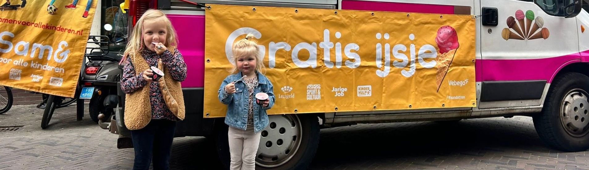 Twee jonge meisjes eten een schepijsje uit een bakje en staan voor een ijscokar voor een banner met de tekst 'gratis ijsje' erop.